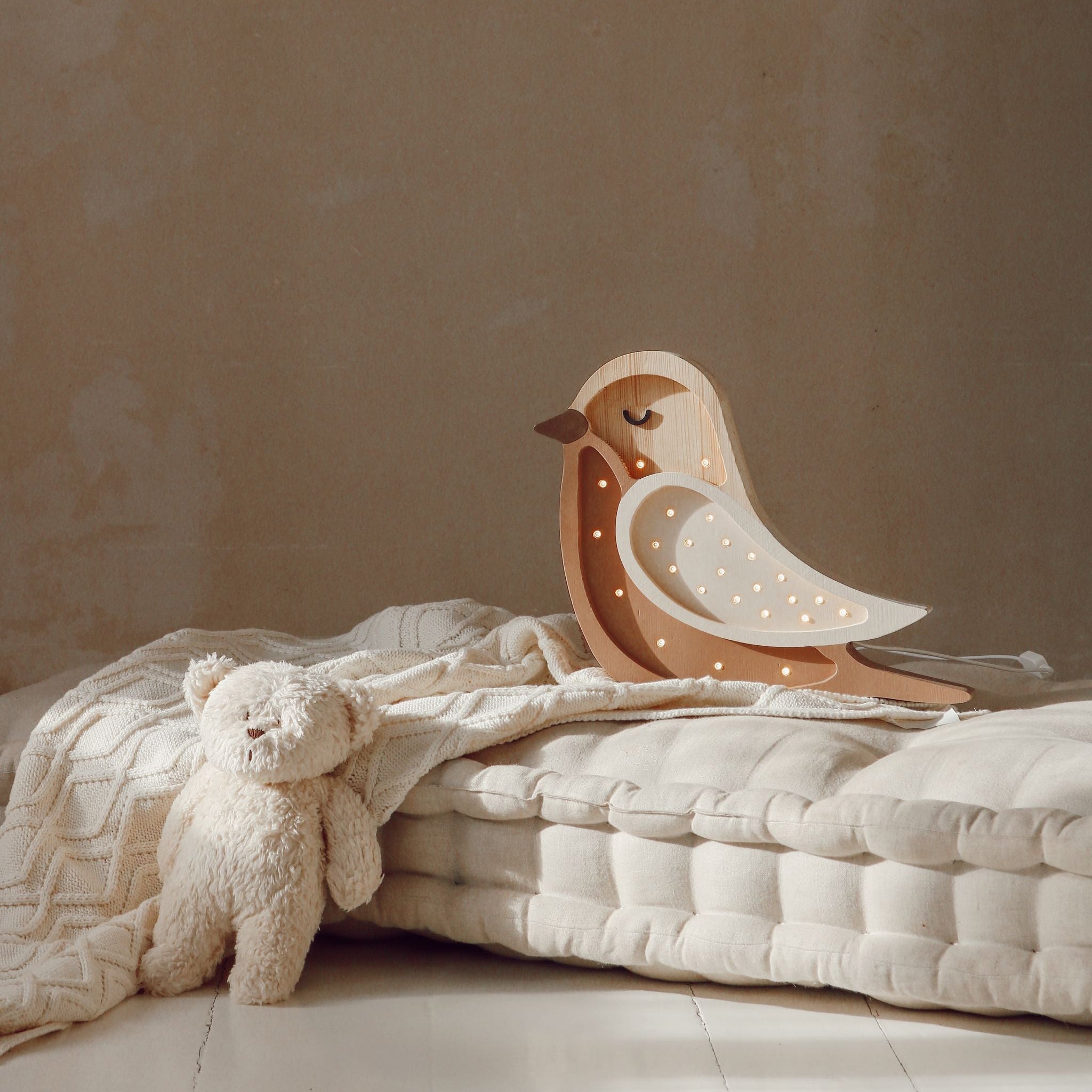 Wooden Little Lights Bird night light on a bed with a soft blanket and teddy bear.