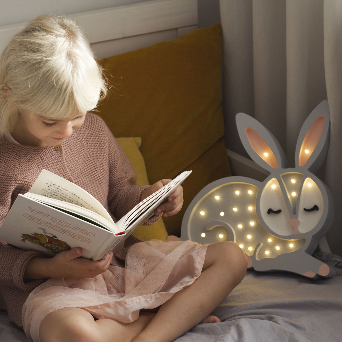 Child reading a book in bed next to Little Lights Bunny night light