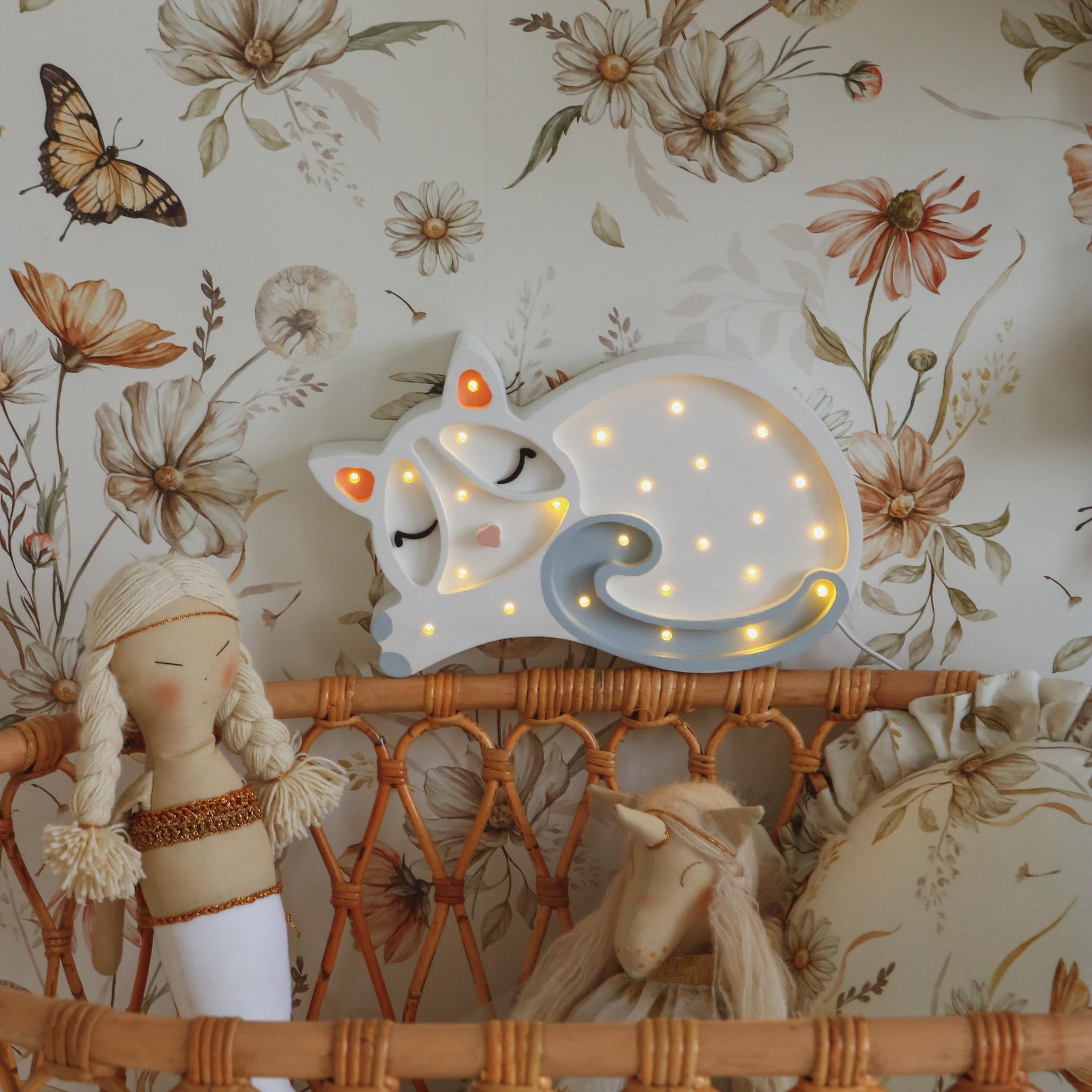 Little Ligts Cat Night Light next to a baby wicker crib against a floral wallpaper