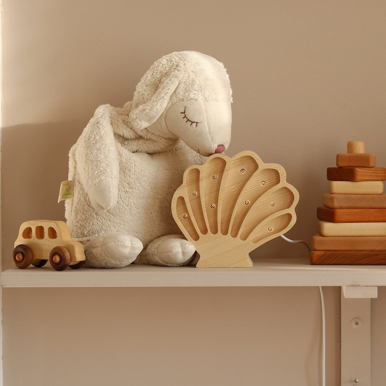A styled collection of wooden and plush toys arranged on a white shelf against a warm beige wall, including a wooden Shell Night Light by Little Lights and a wooden stacking toy.