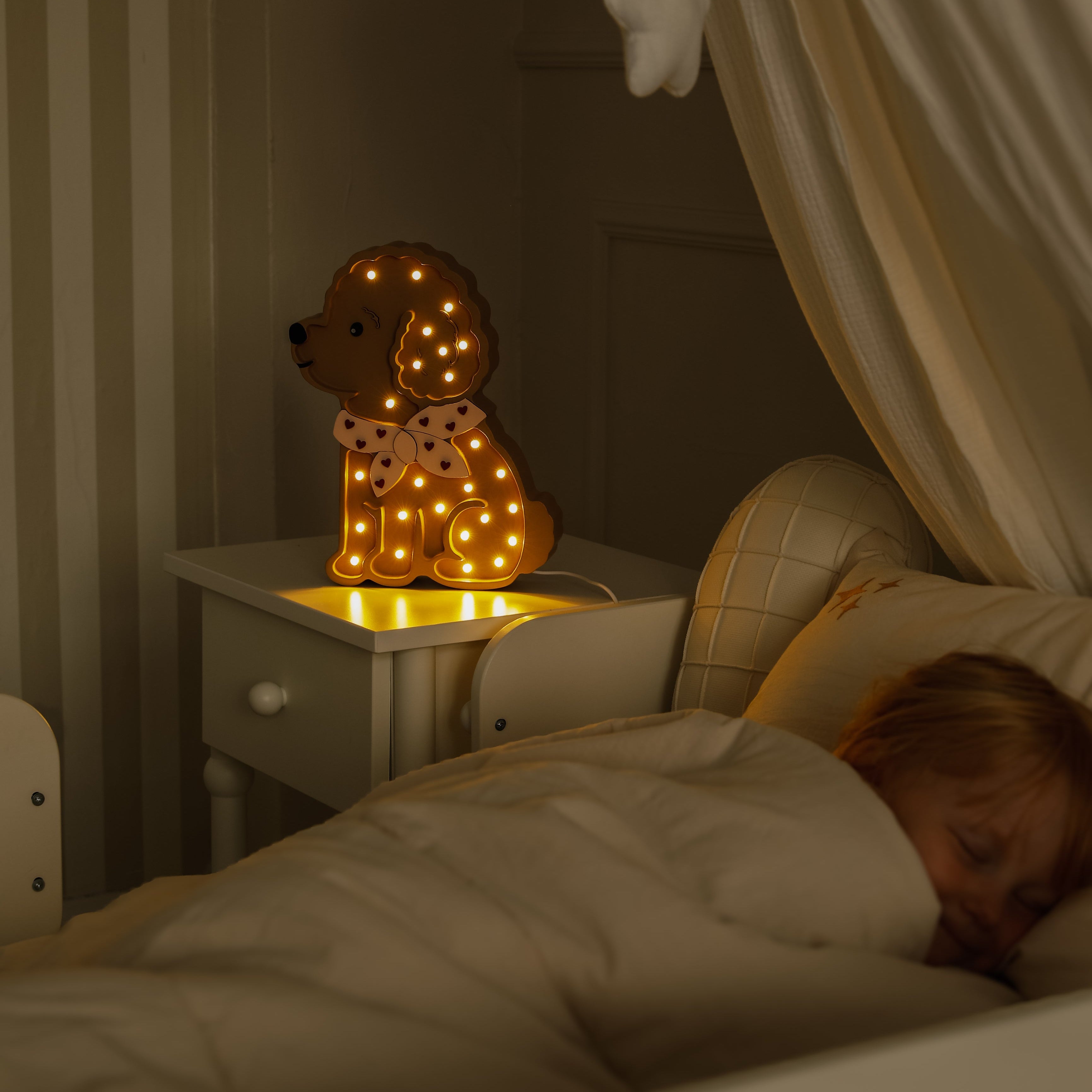 A girl sleeping next to Little Lights Wooden Poodle Dog Lamp glowing softly on the bedside nightstand