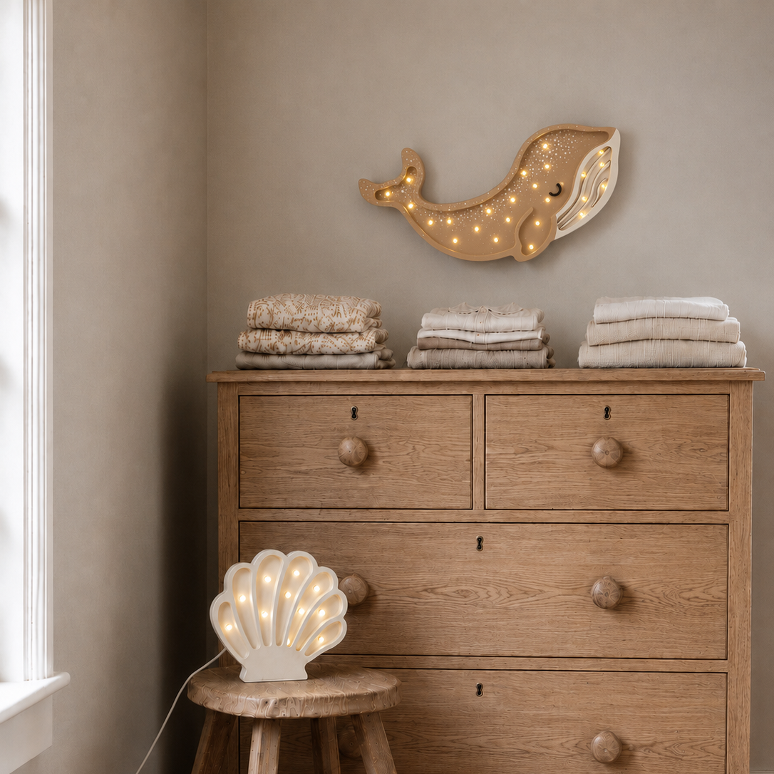 A warm-toned wooden dresser in a styled room corner, with folded textiles on top and a small wooden stool holding a white Shell Lamp. A brown Whale Lamp hangs on the wall above.