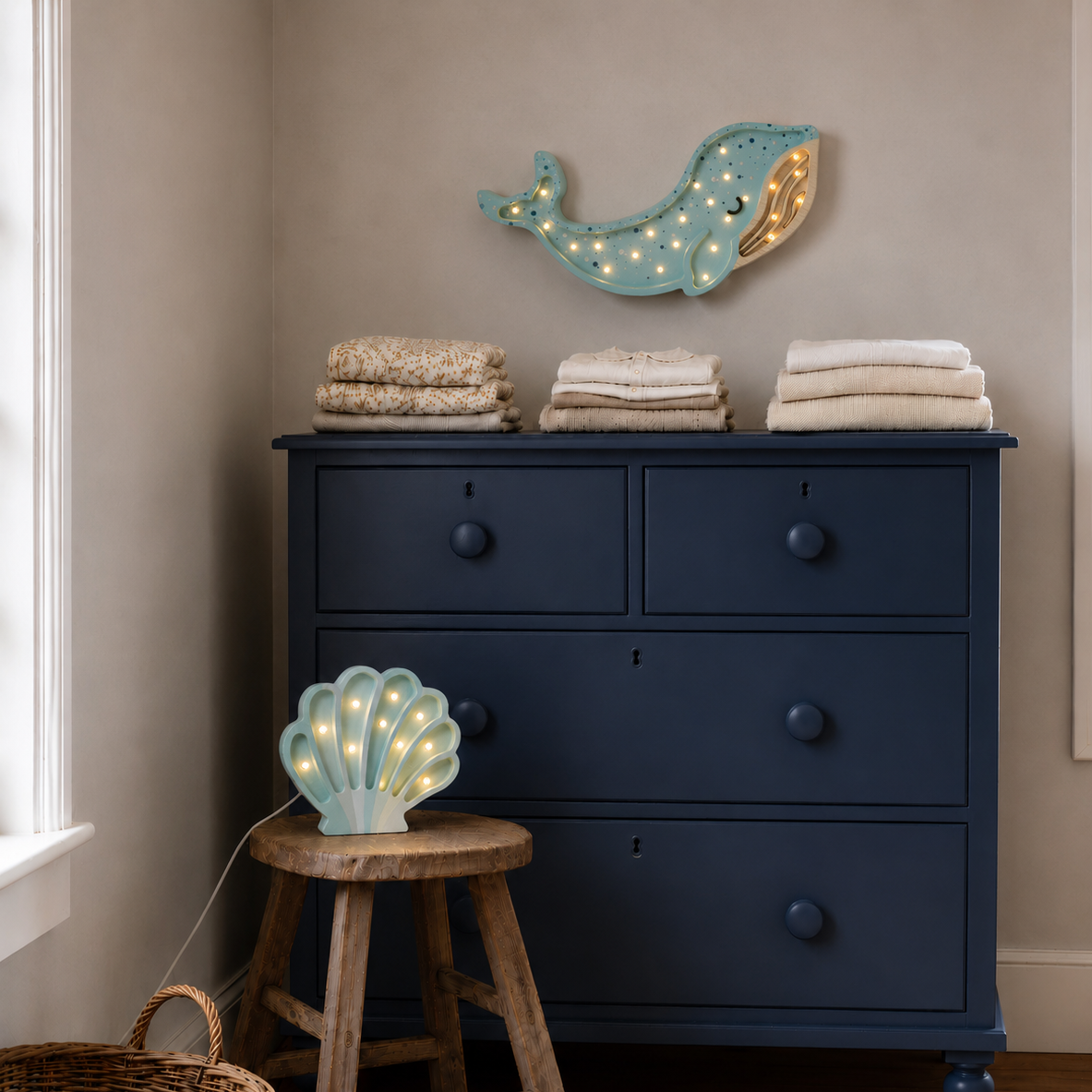 A navy blue wooden dresser with a light blue Whale Lamp hanging above. A small wooden stool holds a blue Shell Lamp by Little Lights.