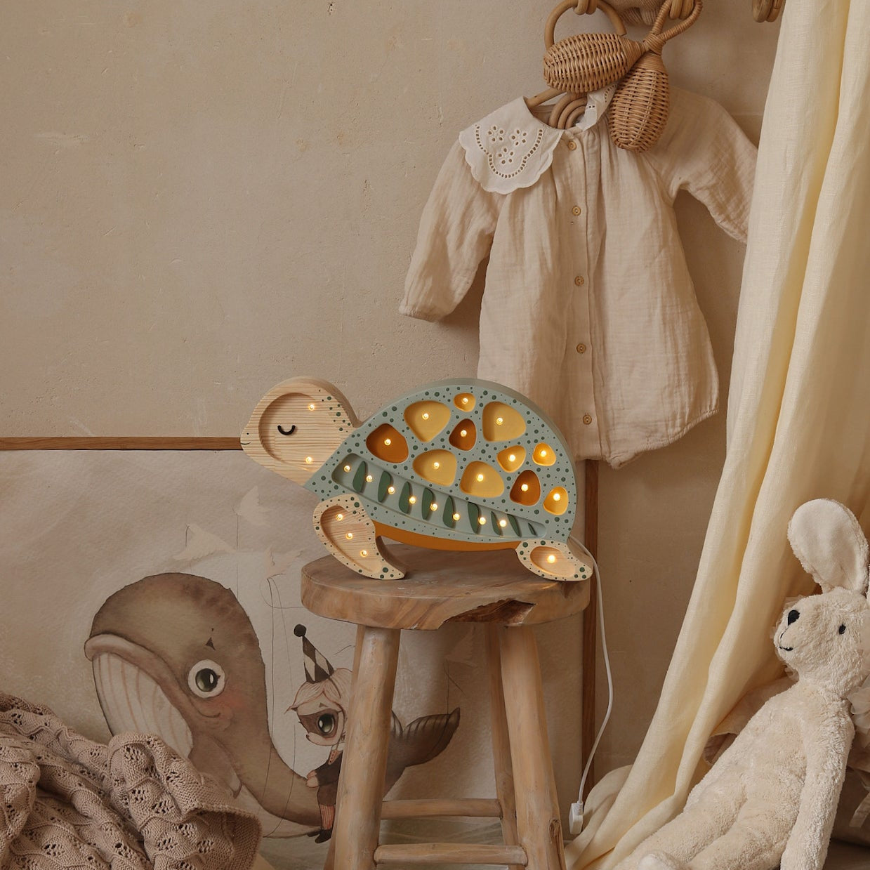 A styled nursery corner featuring a decorative wooden sage turtle night light glowing on a small wooden stool, surrounded by plush toys.